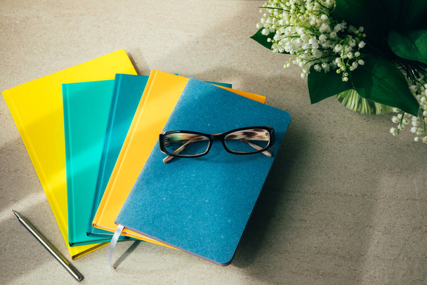Cadernos de escritório multicoloridos sobre um fundo bege claro e óculos no topo com um buquê de lírios do vale. Conjunto de cadernos, vista frontal - Foto, Imagem
