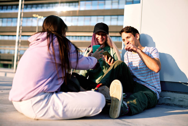 Felices amigos adolescentes están sentados en el exterior urbano y utilizando sus teléfonos para colgar en las redes sociales. - Foto, Imagen