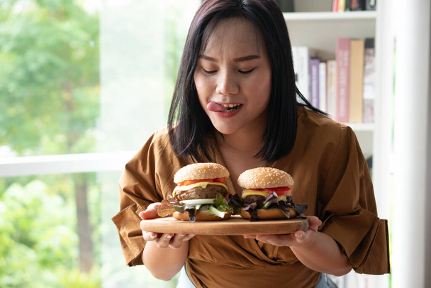 Mujer hambrienta con sobrepeso sosteniendo hamburguesa en un plato de madera, Durante el trabajo desde casa, ganar problema de peso. Concepto de trastorno de atracones (BED)). - Foto, imagen
