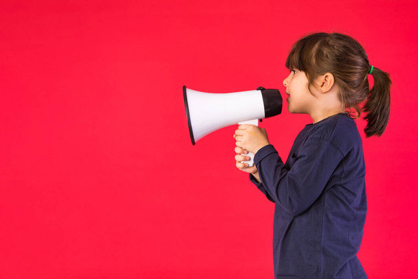 Black-haired girl in a blue sweater and white pants, shouting through a white megaphone, on a red background. - Foto, afbeelding