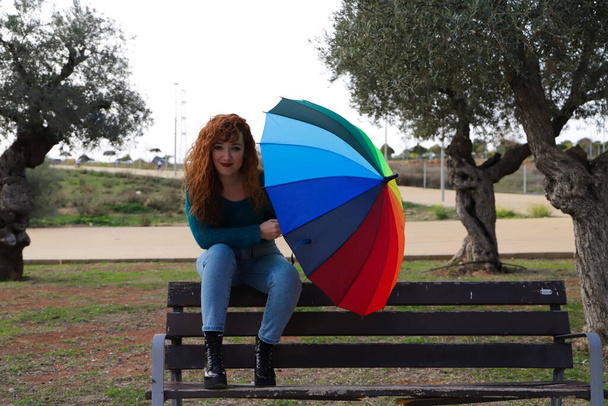 young and beautiful red-haired woman is sitting on a bench in a park where there are many olive trees. She has a colors umbrella to brighten up the grey day. Fashion and beauty concept - Photo, Image