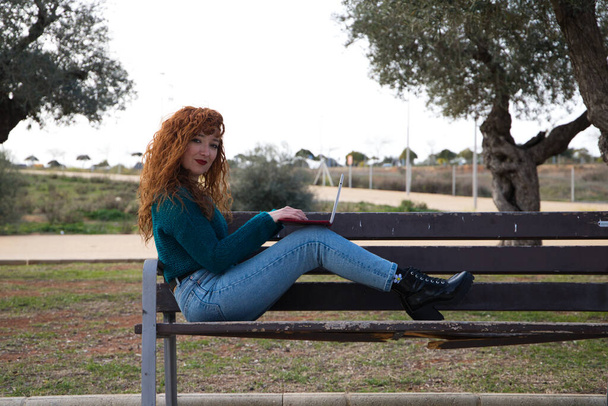 joven y hermosa mujer pelirroja está sentada en un banco en un parque donde hay muchos olivos. Ella está en su computadora portátil revisando las redes sociales y trabajando como una influencia. Concepto de estudiantes. - Foto, imagen