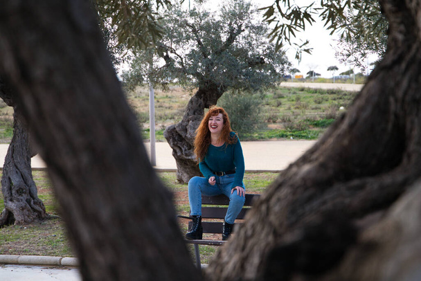 かなり若い赤髪の女性が多くのオリーブの木がある公園のベンチに座っています。撮影されていることに気づかずにっこり笑っている。ファッションと美しさのコンセプト - 写真・画像