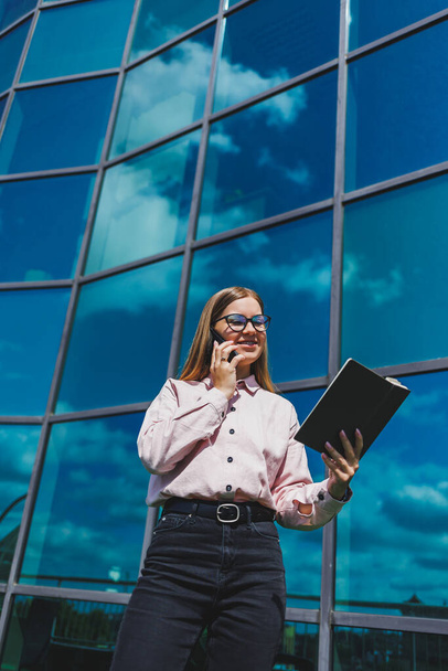 Милая веселая женщина-менеджер в повседневной одежде и модных очках стоит на фоне небоскреба с блокнотом в руках, улыбается и разговаривает по телефону - Фото, изображение