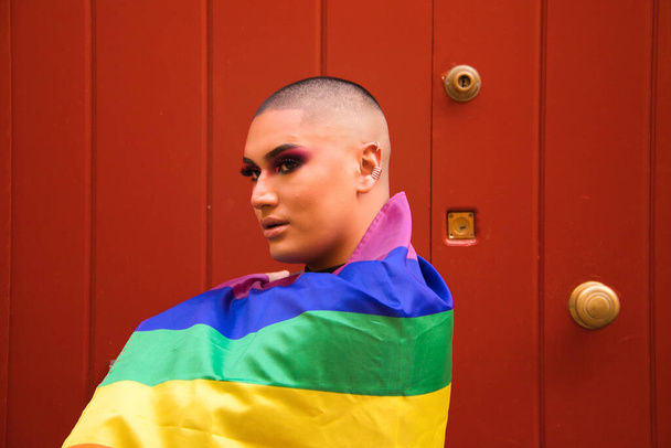 Osoba niebinarna, pochodzenia południowoamerykańskiego i młoda, nosi makijaż i flagę dumy gejowskiej na ramionach. Pojęcie równości, homoseksualizm, gej, lesbijki, duma gejów. - Zdjęcie, obraz