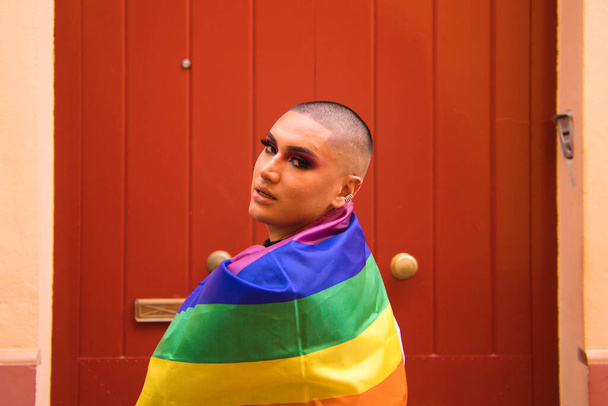 Nicht binäre Person, südamerikanischer Herkunft und jung, die Person trägt Make-up und trägt die Gay Pride Flagge auf den Schultern. Konzept Gleichberechtigung, Homosexualität, schwul, lesbisch, gay pride. - Foto, Bild