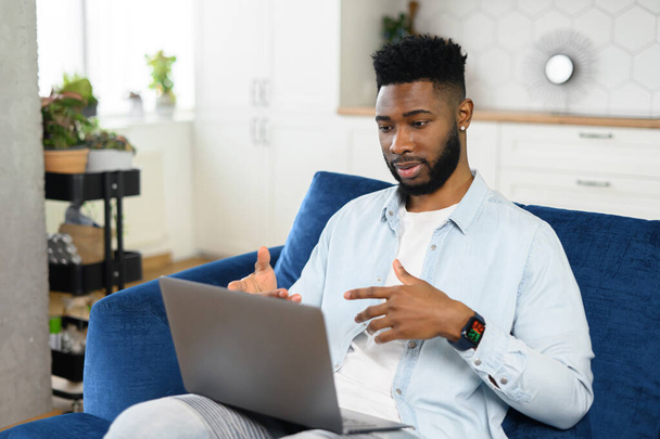 Smiling young bearded man sitting on the couch with laptop on his lap, using dating app, video-calling girlfriend online, gesturing and talking, blogger streaming his daily life - Foto, Imagen
