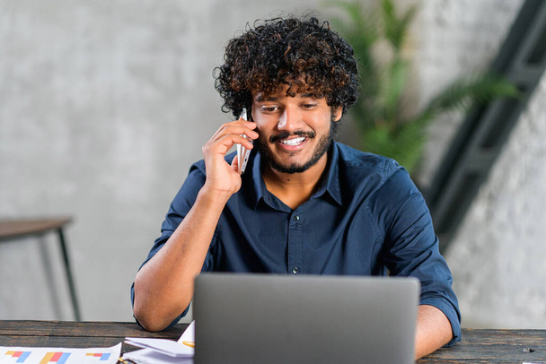 Mid adult Indian man working in his home office, using smartphone and laptop for communication with customers or colleagues. Smiling multiracial eastern guy talking phone watching on computer screen - Foto, afbeelding