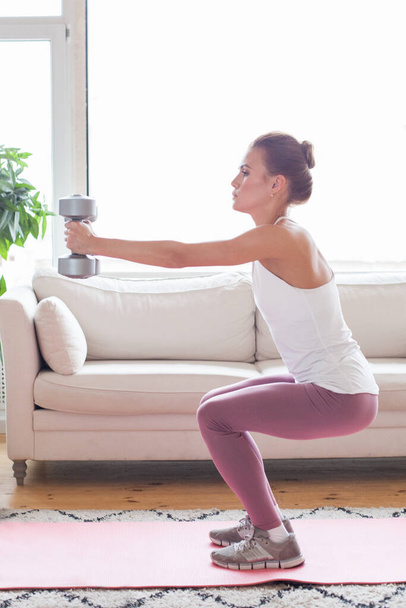 Молодая взрослая женщина делает упражнения с серыми гантелями у себя дома в гостиной - Фото, изображение
