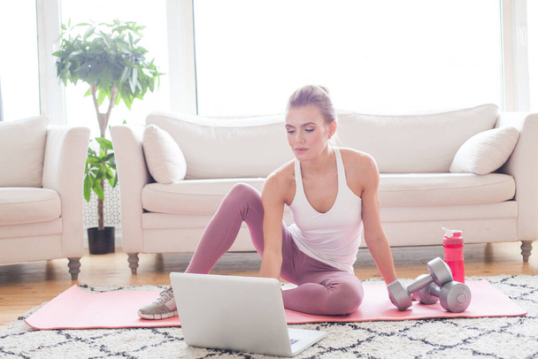 Симпатичная женщина делает упражнения и смотрит онлайн-уроки на ноутбуке, тренируется в гостиной - Фото, изображение
