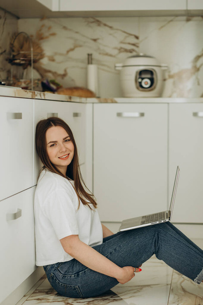 Beautiful woman working with laptop sitting on the floor in the kitchen. - Foto, afbeelding