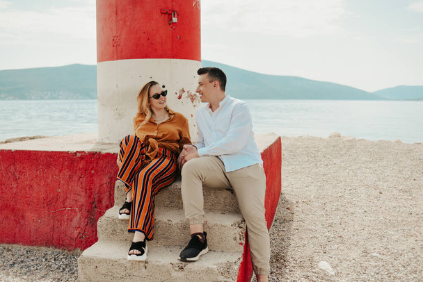 Un couple est assis près d'un phare sur la plage dans le contexte de la mer et des montagnes. Vacances amour, lune de miel, voyage. Couple élégant rire et parler. - Photo, image