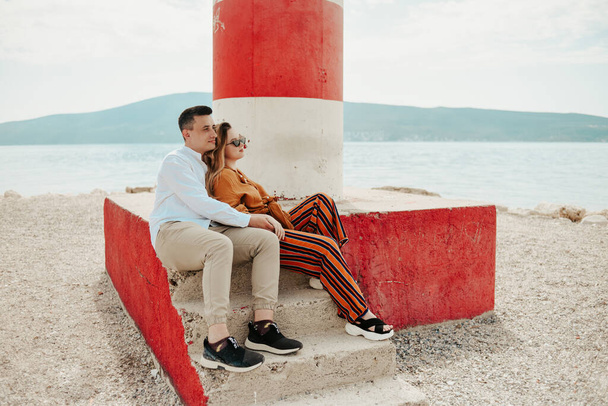 Een paar zit in de buurt van een vuurtoren op het strand tegen de achtergrond van de zee en bergen. Vakantie liefde, huwelijksreis, reizen. stijlvol koppel - Foto, afbeelding