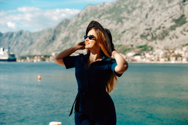 Жінка в сонцезахисних окулярах і капелюх стоїть на тлі моря і гір, посміхаючись. Відпустка та відпочинок на свіжому повітрі. Дівчина з довгим волоссям у відпустці. Мода
 - Фото, зображення