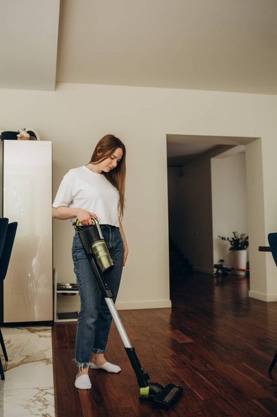 A young Caucasian woman vacuums a kitchen floor using a cordless vertical vacuum cleaner, or electric broom. The maid is cleaning up the hostels cook room. One white female do cleanup of indoors - Photo, image