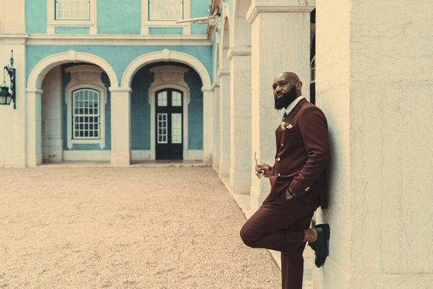A dapper handsome African American bald bearded guy in a dark-red tailored costume is leaning against the wall, looking away and holding eyeglasses in his hand; a copy space place on the left - Foto, Bild