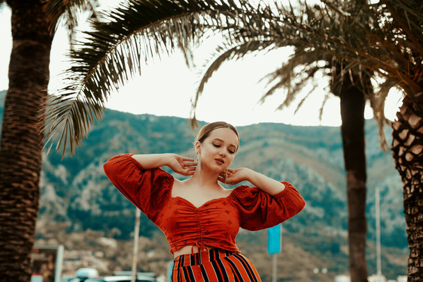Stijlvolle vrouw in de zomer op de achtergrond van palmbomen en bergen. Spaans meisje in een rood pak en lange oorbellen - Foto, afbeelding