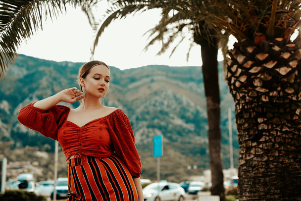 Stilvolle Frau im Sommer vor dem Hintergrund von Palmen und Bergen. Spanierin in rotem Anzug und langen Ohrringen - Foto, Bild