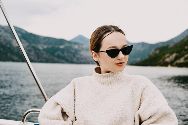 A girl in sunglasses and a white suit rides on a yacht. Journey to the mountains by sea. Traveling - Zdjęcie, obraz
