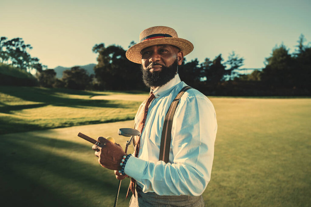 A portrait of a dashing mature bearded black man in a fashionable outfit with a straw hat and braces standing on a golf field in the evening with a sunset, holding a golf club and a cigar in the hands - Photo, Image
