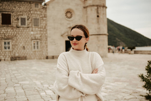 Een meisje met een zonnebril en een wit pak loopt op het eiland. Reizen, Montenegro. Boka eiland Kerk van Onze Lieve Vrouw van de Rotsen Kotor Bay - Foto, afbeelding