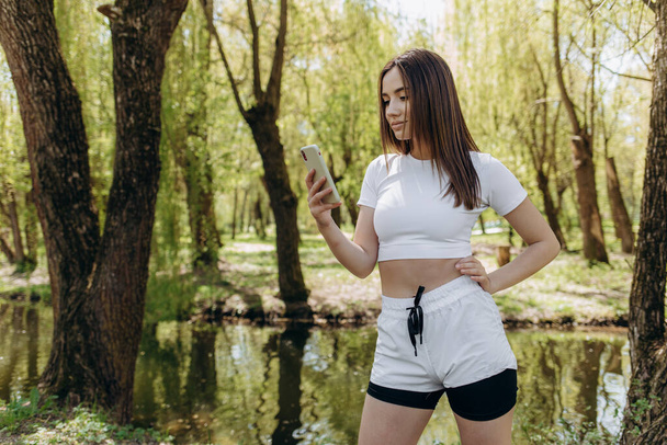 Καυκάσια κοπέλα με αθλητική στολή χαμογελάει και χρησιμοποιεί το κινητό της σε ένα πράσινο πάρκο.. - Φωτογραφία, εικόνα