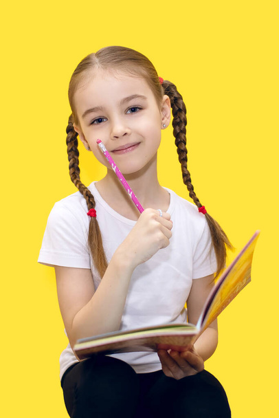 A cute little girl is learning lessons while holding a notebook and a pencil in her hand, or drawing while sitting on a yellow background. Child with pigtails is thoughtful and smiling - Photo, Image