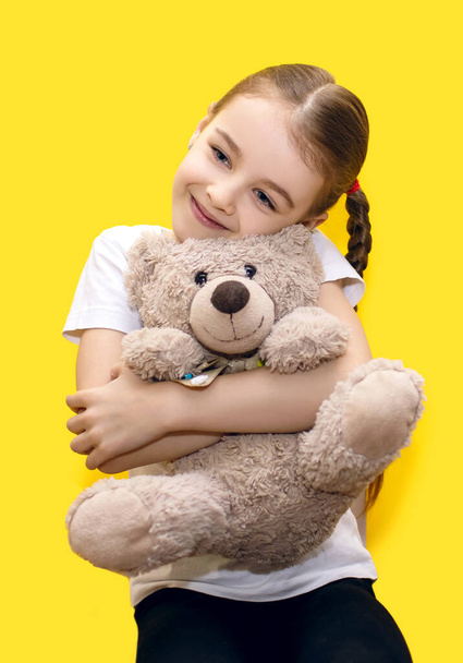 Cute little girl hugging a teddy bear while sitting on a yellow background. A beautiful preschool girl with pigtails smiles and looks good-natured. Happy girl with her favorite toy - Fotografie, Obrázek