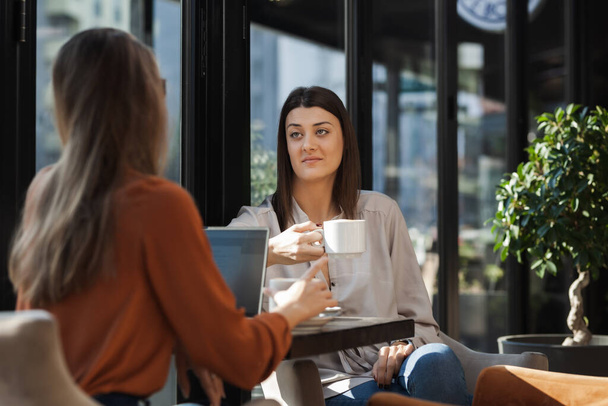 Two young business women in a cafe having one on one meeting. Friends after work talking gossiping and having coffee at a window table on a sunny day. - Fotografie, Obrázek