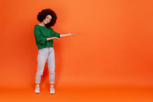 Full length portrait of smiling woman with Afro hairstyle wearing green casual style sweater presenting copy space for promotional text. Indoor studio shot isolated on orange background. - Photo, image