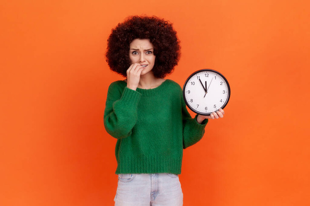 Portrait of nervous woman with Afro hairstyle wearing green casual style sweater holding wall clock and biting fingernails, worried of deadline. Indoor studio shot isolated on orange background. - Foto, afbeelding