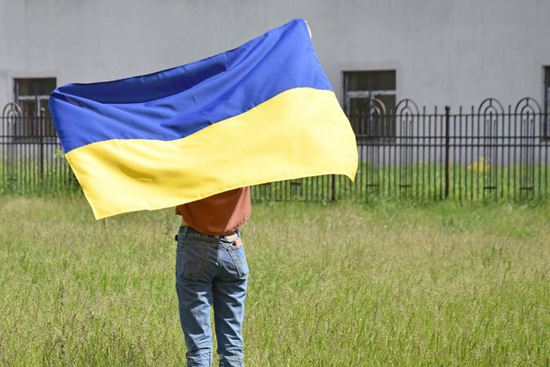地元の公園を背景に、ウクライナの国旗で覆われた若い女の子が立っています。青と黄色の色。ウクライナでの戦争. - 写真・画像