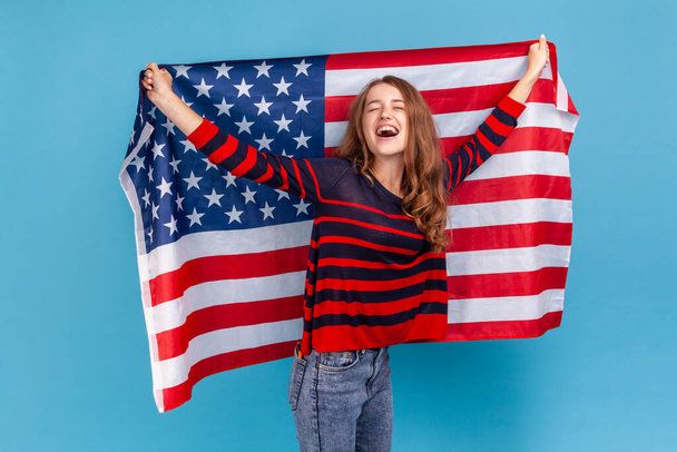 Excited young adult woman wearing striped casual style sweater, holding huge american flag and yelling happily, celebrating national holiday. Indoor studio shot isolated on blue background. - Photo, image
