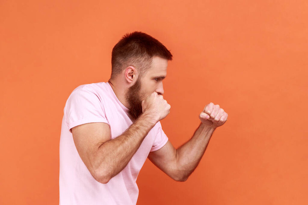 Side view of bearded man aggressive fighter holding clenched fists up ready to boxing, martial art trainer, self defense, wearing pink T-shirt. Indoor studio shot isolated on orange background. - Fotoğraf, Görsel