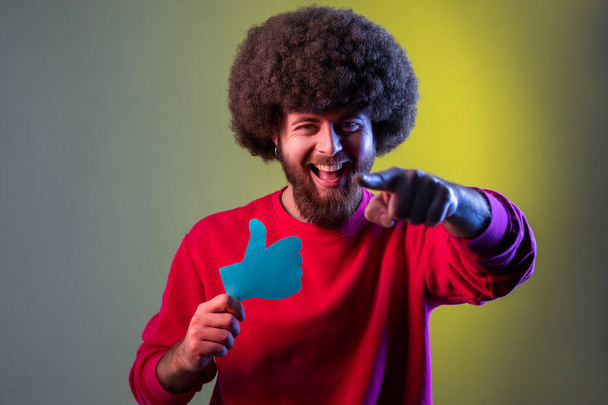 Portrait of excited hipster man with Afro hairstyle holding and showing like or thumbs up paper shape sign, pointing finger to camera. Indoor studio shot isolated on colorful neon light background. - Foto, imagen