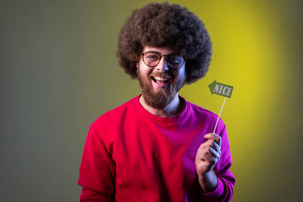 Happy positive hipster man with Afro hairstyle having event, holding party props with nice word in hands, wearing red sweatshirt. Indoor studio shot isolated on colorful neon light background. - Foto, Bild