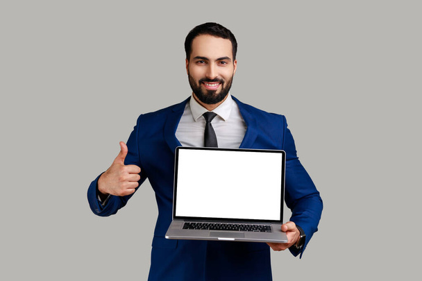 Portrait of satisfied delighted bearded man working on laptop with smile and showing thumb up, looking at camera, wearing official style suit. Indoor studio shot isolated on gray background. - Photo, image
