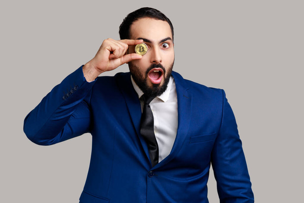 Portrait of bearded businessman covering eye with golden bitcoin, looking at camera with shocked expression, wearing official style suit. Indoor studio shot isolated on gray background. - Фото, изображение