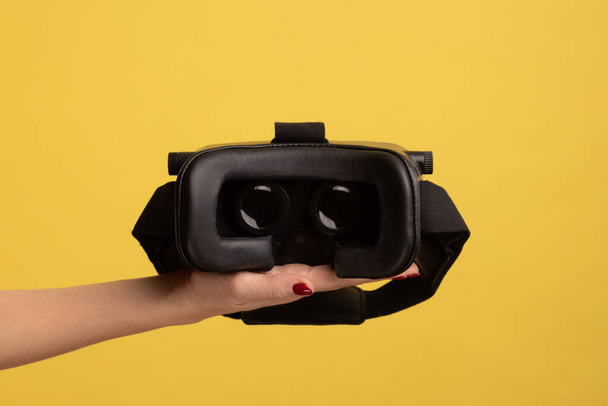 Side view of woman hand holding VR goggles, virtual reality 3d glasses headset for cyberspace simulation, innovative technologies, future and science. Indoor studio shot isolated on yellow background. - Φωτογραφία, εικόνα