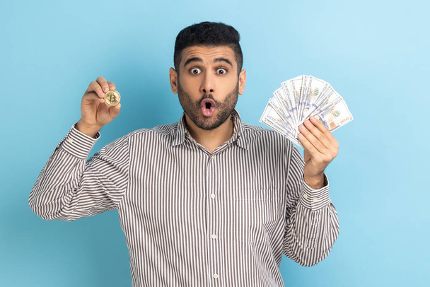 Shocked businessman holding showing dollar bills and golden coin of crypto currency, transferring money into digital bitcoins, wearing striped shirt. Indoor studio shot isolated on blue background. - Фото, изображение