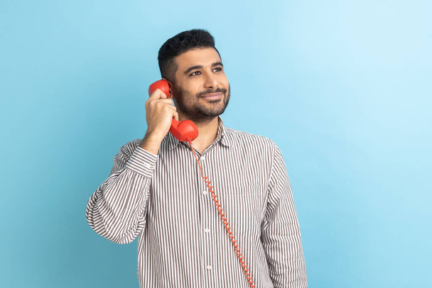 Positive happy businessman with beard talking landline telephone holding in hand handset, looking at camera with toothy smile, wearing striped shirt. Indoor studio shot isolated on blue background. - Fotoğraf, Görsel
