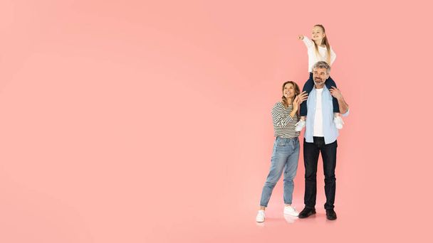 Positieve familie van drie poseren, dochter wijzende vinger op lege ruimte zitten op papa 's schouders poseren over Pink Studio achtergrond. Ouders en reclame voor kinderen. Panorama - Foto, afbeelding