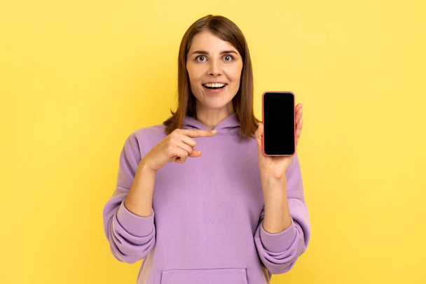 Positiv entzückte Frau zeigt auf Smartphone mit leerem Display für Werbetext, blickt lächelnd in die Kamera, trägt lila Kapuzenpulli. Indoor Studio aufgenommen isoliert auf gelbem Hintergrund. - Foto, Bild