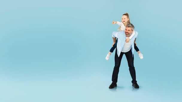 Happy Middle Aged Father Carrying Little Daughter Piggyback Pointing Finger Showing Free Space For Text Advertising Great Offer Posing Over Blue Studio Background. Panorama, Full Length - Fotó, kép
