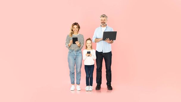 Cheerful Family Using Smartphone, Laptop And Digital Tablet Computers Browsing Internet Via Gadgets Smiling To Camera Standing Over Pink Studio Background. Panorama, Full Length - Фото, изображение