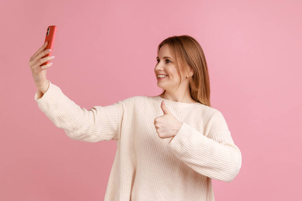 Portrait of happy blond woman blogger broadcasting livestream, showing thumb up to mobile phone camera, expressing positive, wearing white sweater. Indoor studio shot isolated on pink background. - Φωτογραφία, εικόνα