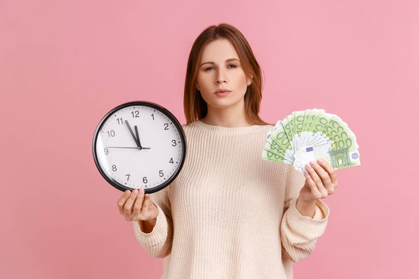 Portrait of serious strict blond woman holding big wall clock and euro banknotes, looking at camera with bossy expression, wearing white sweater. Indoor studio shot isolated on pink background. - Foto, imagen