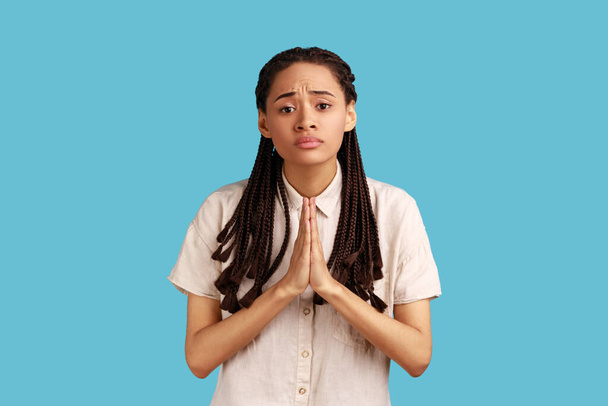 Cute woman with dreadlocks keeps palms pressed together, pleading angel, has innocent expressions, asks for mercy or apologize, wearing white shirt. Indoor studio shot isolated on blue background. - Fotografie, Obrázek