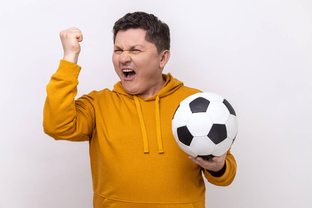 Positive satisfied man screaming holding soccer ball, celebrating victory of favourite football team on championship, wearing urban style hoodie. Indoor studio shot isolated on white background. - Foto, Bild