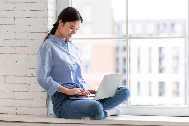 Kaunis nuori aasialainen nainen sinisessä paidassa ja farkut istuu ikkunalaudalla kotona mukavalla kaupunkinäkymällä, käyttäen kannettavaa tietokonetta, nainen freelancer työskentelee verkossa, kirjoittamalla tietokoneen näppäimistöllä, panoraama kopioi tilaa - Valokuva, kuva